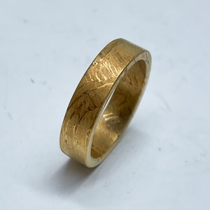Könnte beinhalten: Ein goldfarbener Ring mit einem rechteckigen Band. Der Ring hat eine strukturierte Oberfläche mit sichtbaren Linien und Unvollkommenheiten. Der Ring ist wahrscheinlich aus Metall und ist zum Tragen am Finger bestimmt.