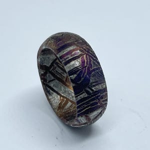 Puede incluir: Primer plano de un anillo estampado con una mezcla de tonos plateados, morados y marrones. El anillo tiene un diseño texturizado único con líneas y patrones visibles. El anillo es de estilo banda.