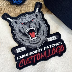 May include: Embroidered patch featuring a snarling bear with red eyes and hockey sticks. The patch is gray, black, and red, with the text "EMBROIDERY PATCHES CUSTOM LOGO". The patch is likely for apparel or accessories.