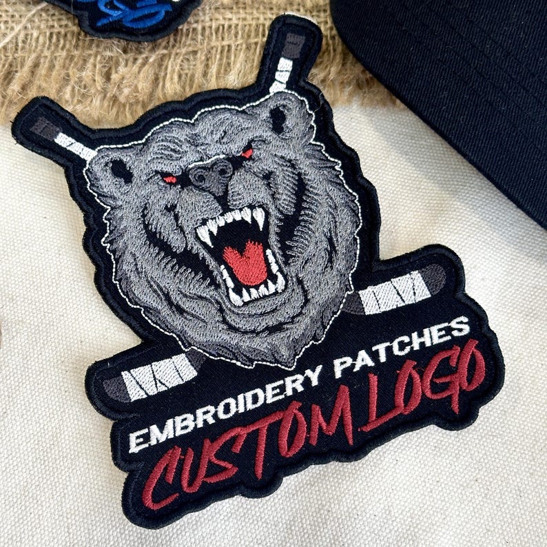 May include: Embroidered patch featuring a snarling bear with red eyes and hockey sticks. The patch is gray, black, and red, with the text "EMBROIDERY PATCHES CUSTOM LOGO". The patch is likely for apparel or accessories.