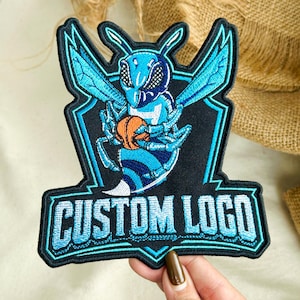 May include: An embroidered patch featuring a stylized blue hornet mascot holding an orange basketball, centered within a black shield-shaped border. The text CUSTOM LOGO is stitched in light blue block letters at the bottom of the patch design.