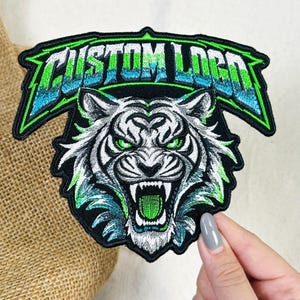 May include: An embroidered patch featuring a snarling white tiger head with bright green eyes and mouth. Above the tiger, a banner displays the text CUSTOM LOGO in a stylized font with a gradient of green, white, and blue thread colors.