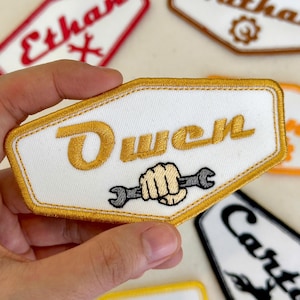 May include: Embroidered patch with the name "Owen" in gold lettering on a white background. The patch features a gold border and a graphic of a fist holding a wrench. Other patches with names are visible.