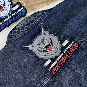May include: Embroidered patches featuring a snarling bear with hockey sticks and the text "CUSTOM LOGO." The patches are gray, red, and black, and are displayed on a denim jacket.