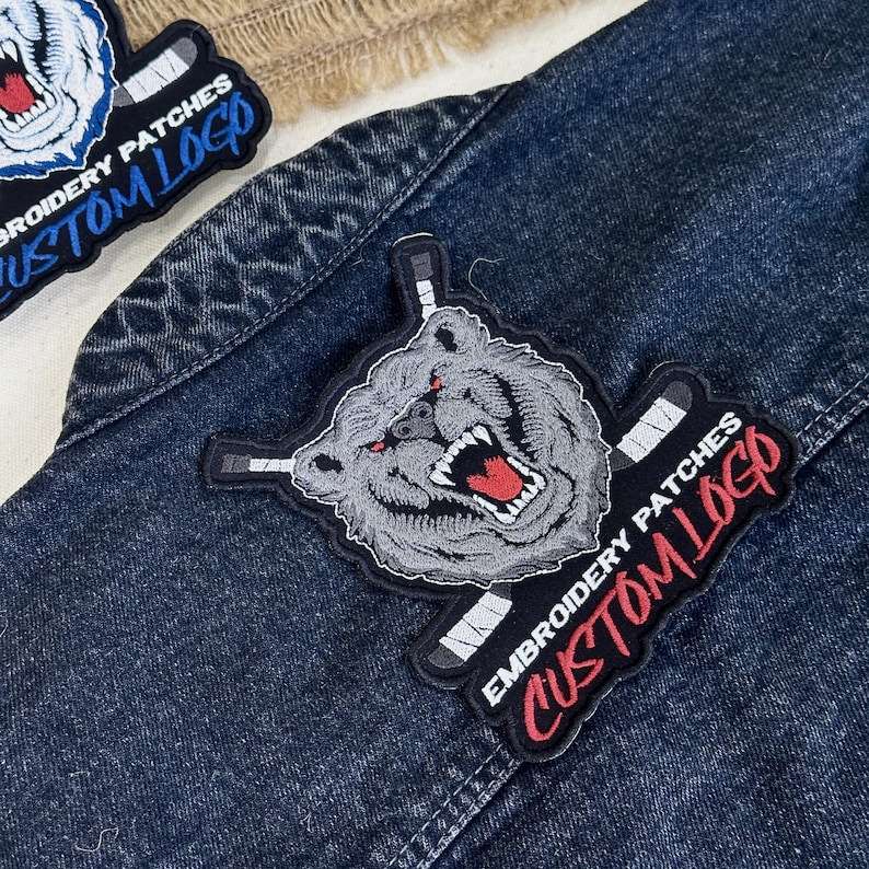 May include: Embroidered patches featuring a snarling bear with hockey sticks and the text "CUSTOM LOGO." The patches are gray, red, and black, and are displayed on a denim jacket.