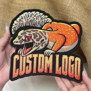 May include: An embroidered patch featuring a lizard with a brown and tan patterned head, a bright orange body, and a large crest. The lizard has a red eye and an open mouth. Below the lizard, the text CUSTOM LOGO is stitched in a bold, orange-to-red gradient font.