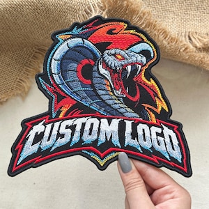 May include: An embroidered patch featuring a cobra with a flared hood, red eyes, and sharp fangs. The design includes vibrant red, orange, and blue flames surrounding the snake. Below the cobra, the text CUSTOM LOGO is stitched in bold letters.