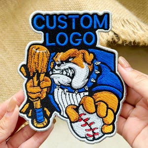 May include: An embroidered patch featuring a bulldog mascot holding a wooden baseball bat and a baseball. The text CUSTOM LOGO is stitched in blue at the top. The design uses black, blue, white, and tan thread with a white border on a black background.