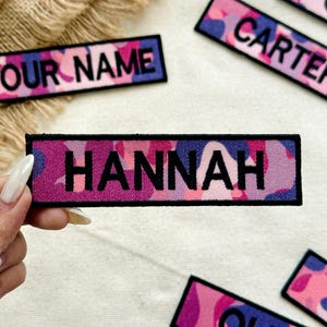 May include: Rectangular patch with the name "HANNAH" embroidered in black on a pink, purple, and blue camouflage pattern. The patch is held by a hand, with other similar patches visible.