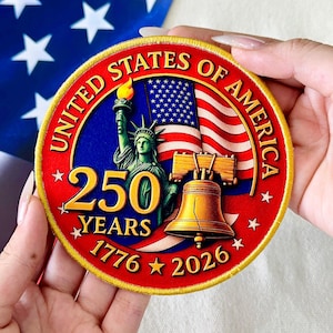 May include: A round, embroidered patch with the text "UNITED STATES OF AMERICA" in gold lettering. The patch features the Statue of Liberty, a waving American flag, and the Liberty Bell. The text "250 YEARS 1776 2026" is also included.