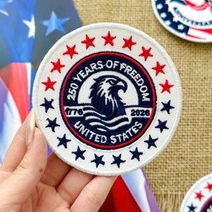 May include: A round white patch with red and blue accents. The patch features red stars around the edge and blue stars. The center design includes an eagle and the text "250 YEARS OF FREEDOM UNITED STATES 1776-2026".