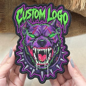May include: An embroidered patch featuring a snarling purple dog with glowing green eyes and sharp teeth, wearing a spiked collar. The text CUSTOM LOGO is stitched in a dripping green font at the top against a black background with purple borders.