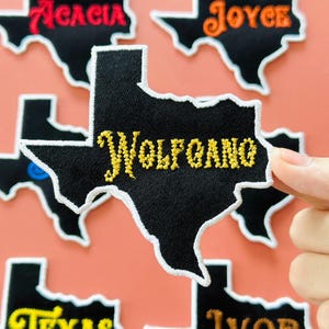 May include: Black Texas-shaped embroidered patches with white borders and various names in colorful thread. One patch held by a hand reads "Wolfgang" in yellow script. Other patches include names like "Acacia" and "Joyce".