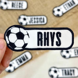 May include: White and black soccer-themed name patches. The patches have a soccer ball design on the left and a name in bold black letters on a white background. The name "Rhys" is featured prominently, with other names visible.