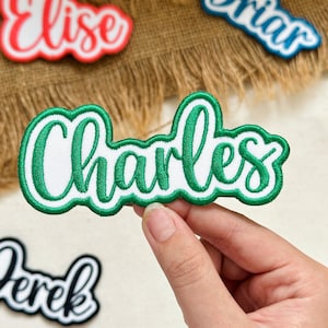 May include: An embroidered name patch featuring the name Charles in a green cursive script font with a white background and a thick green border. The patch has a scalloped edge design and is made of fabric with visible stitching.