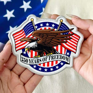 May include: Embroidered patch featuring a brown eagle with a white head, set against a backdrop of the American flag. The patch includes the text "250 YEARS OF FREEDOM" on a banner, with red, white, and blue colors.