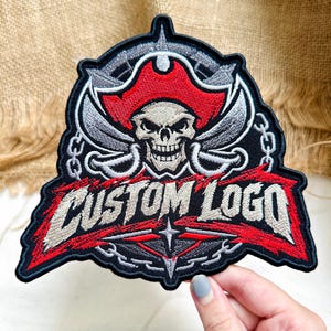 May include: An embroidered patch featuring a skull wearing a red pirate hat, positioned above two crossed silver swords and a chain border. The text CUSTOM LOGO is stitched in silver thread across a red background at the bottom of the patch.