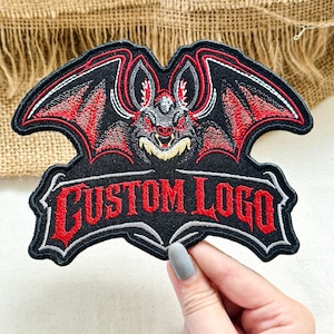 May include: An embroidered patch featuring a central bat with red and black wings, pointed ears, and a cream-colored chest. Below the bat, the text CUSTOM LOGO is stitched in bold red lettering with a black outline on a black fabric background.