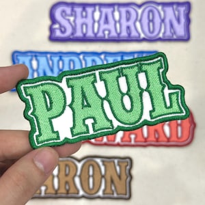 May include: Embroidered name patches in various colors. The patch held in the hand says "PAUL" in green with a white border. Other patches include the names "SHARON", "LINDA", and "RON". The patches have a retro, varsity style.