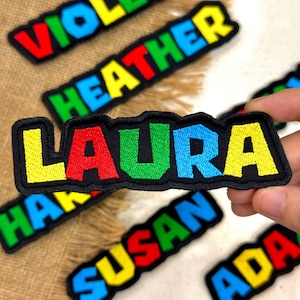 May include: Embroidered name patches in various colors. The name "LAURA" is in the foreground, with red, green, blue, and yellow letters on a black background. Other names like "HEATHER" and "SUSAN" are also visible.