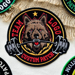 Pu&ograve; includere: Toppa ricamata con un lupo marrone con occhi rossi, che tiene un bilanciere in bocca. La toppa ha uno sfondo nero con le parole "TEAM LOGO CUSTOM PATCH" in rosso. Una stella d'oro in alto.