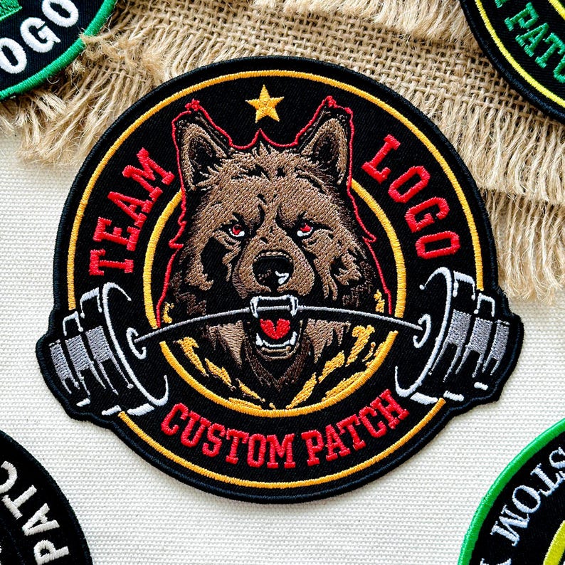 Pu&ograve; includere: Toppa ricamata con un lupo marrone con occhi rossi, che tiene un bilanciere in bocca. La toppa ha uno sfondo nero con le parole "TEAM LOGO CUSTOM PATCH" in rosso. Una stella d'oro in alto.