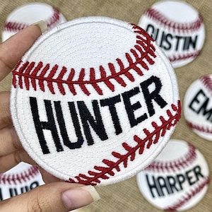 May include: White baseball patch with red stitching and the name "HUNTER" in black. Additional patches with names "AUSTIN", "EM", and "HARPER" are visible. The patches are round and resemble baseballs.