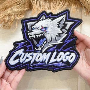 May include: An embroidered patch featuring a snarling wolf head in shades of gray, white, and blue, set against a black background with purple accents. The text CUSTOM LOGO is stitched in white with a bold, slanted font across the lower section.