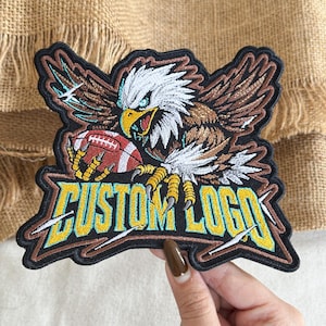 May include: An embroidered patch featuring a bald eagle with white, brown, and black feathers, holding a red and white football in its yellow talons. Below the eagle, the text CUSTOM LOGO is stitched in bold yellow letters with a light blue outline.