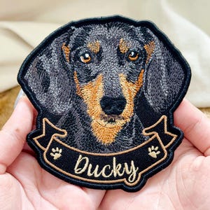 May include: An embroidered patch featuring a detailed portrait of a black and tan dachshund dog. Below the dog's head is a black banner with gold trim and gold paw prints on each side. The text Ducky is embroidered in gold script on the banner.