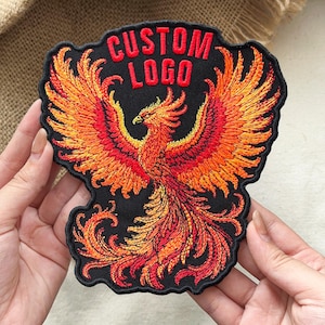 May include: A black embroidered patch featuring a phoenix with vibrant red, orange, and yellow wings and tail feathers. The text CUSTOM LOGO is stitched in bold red thread at the top of the patch, which has a black border and a die-cut shape.
