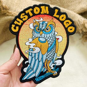 May include: An embroidered patch featuring a central owl illustration in shades of blue, yellow, and white against a circular orange sun background. The black border contains the yellow text CUSTOM LOGO arched across the top of the design.