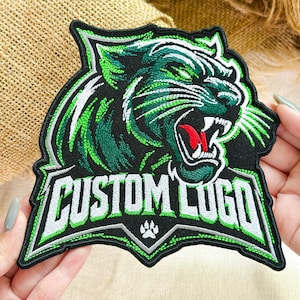 May include: An embroidered patch featuring a stylized panther head in black, green, and white thread. The panther has a red tongue and bared teeth. Below the panther, the text CUSTOM LOGO is stitched in white with green outlines, with a small paw print.