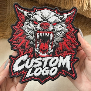 May include: An embroidered patch featuring a snarling wolf head with white fur, red eyes, and a red mouth with sharp teeth. The design includes a black background with red jagged borders and the white text CUSTOM LOGO at the bottom.