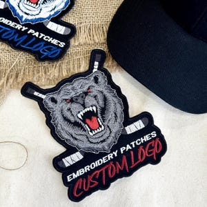 May include: Embroidered patches featuring a snarling bear with red eyes and hockey sticks. The patches have the text "EMBROIDERY PATCHES CUSTOM LOGO" in white and red. A black baseball cap is in the background.