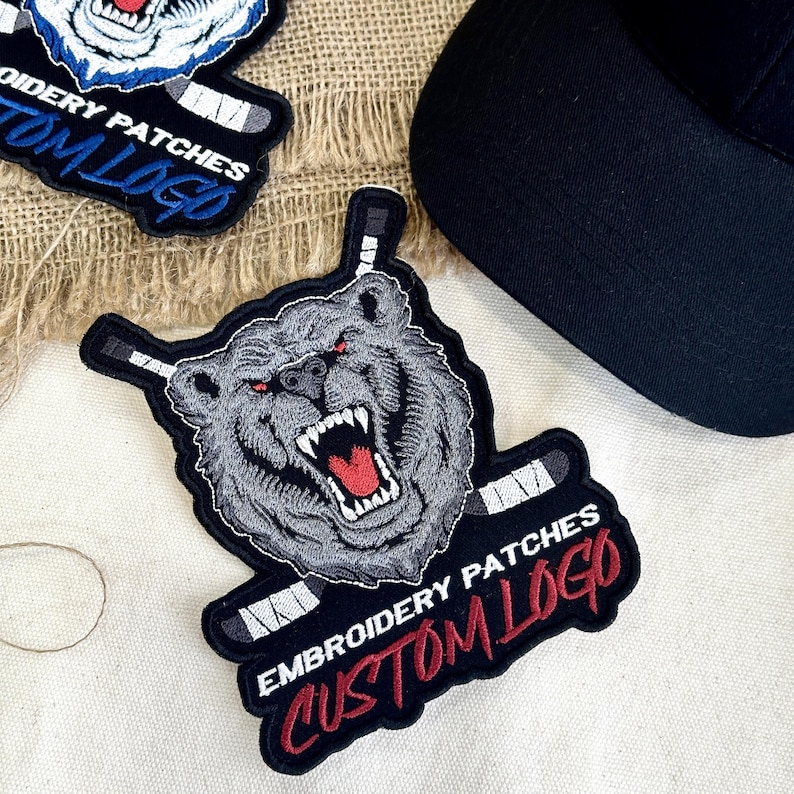 May include: Embroidered patches featuring a snarling bear with red eyes and hockey sticks. The patches have the text "EMBROIDERY PATCHES CUSTOM LOGO" in white and red. A black baseball cap is in the background.