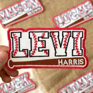 May include: Embroidered baseball-themed patch with the name "LEVI HARRIS" in white letters with red stitching, set against a baseball bat design. The patch has a red border and is surrounded by other name patches.