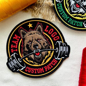 Pu&ograve; includere: Toppa ricamata con un lupo marrone con occhi rossi che tiene un bilanciere. Sfondo nero con dettagli gialli e rossi. Testo: "TEAM LOGO CUSTOM PATCH".