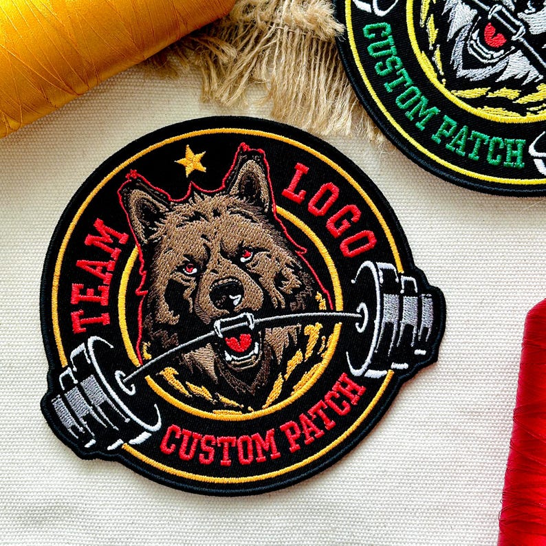 Pu&ograve; includere: Toppa ricamata con un lupo marrone con occhi rossi che tiene un bilanciere. Sfondo nero con dettagli gialli e rossi. Testo: "TEAM LOGO CUSTOM PATCH".
