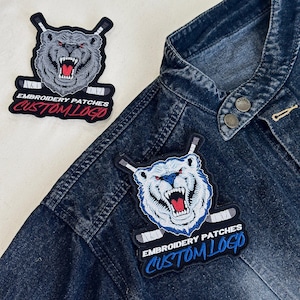 May include: Embroidered patches featuring a snarling bear with hockey sticks. The patches have the text "EMBROIDERY PATCHES CUSTOM LOGO" in white and red. One patch is on a denim jacket.