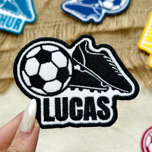 May include: Embroidered patch featuring a soccer ball and a soccer cleat in black and white. The patch has a white border and the name "LUCAS" in bold, white letters. Other patches are visible in the background.