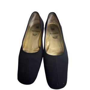 May include: A pair of black slip-on flats with a square toe design. The interior features a gold-colored lining with the text Bruno Magli, Made in Italy, Bologna printed on the insole. The exterior is a solid black fabric with a center seam.