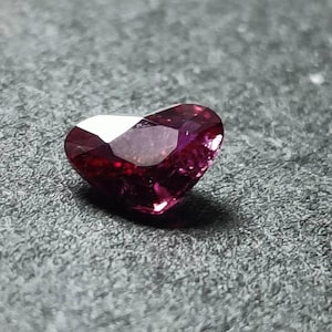 May include: A heart-shaped gemstone with a deep magenta and dark red hue. The stone features multiple faceted surfaces that reflect light, creating a brilliant appearance with sharp edges and a pointed base, showcasing a clear, translucent quality.