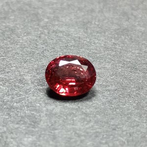 May include: A loose, oval-cut gemstone with a vibrant, deep red colour and a faceted surface that reflects light. The stone displays a clear, transparent quality with multiple geometric facets across its top, creating a brilliant, sparkling appearance.