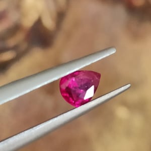 以下が含まれることがあります： 銀色のピンセットの2つの金属製の先端の間にしっかりと保持された、鮮やかで深いピンクがかった赤色の洋ナシ型のファセットカットされた宝石。石は内部の光の反射と滑らかで磨かれた表面の質感を示しています。