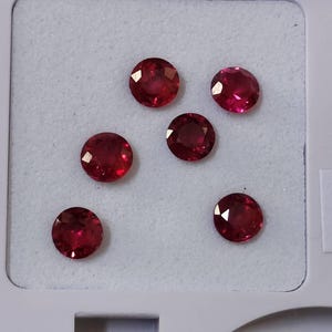 Könnte beinhalten: Fünf runde, facettierte, tiefrote Edelsteine, die in einer losen Anordnung in einem weißen Etui liegen. Jeder Stein weist einen Brillantschliff mit zahlreichen dreieckigen Facetten auf, die das Licht reflektieren und eine lebendige, gesättigte karmesinrote Farbe zeigen.