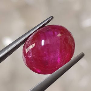以下が含まれることがあります： 鮮やかなピンクがかった赤色をした、楕円形のカボションカットの宝石。表面は滑らかに磨かれており、光を反射して内部の透明感や結晶構造に見られる自然な模様を映し出しています。宝石は金属製のピンセットで挟まれています。