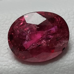 Könnte beinhalten: Ein oval geschliffener Edelstein in einer tiefen, leuchtenden magentaroten Farbe. Der Stein weist eine komplexe innere Struktur mit sichtbaren Einschlüssen und lichtreflektierenden Facetten auf der Oberfläche auf, die für Tiefe sorgen.