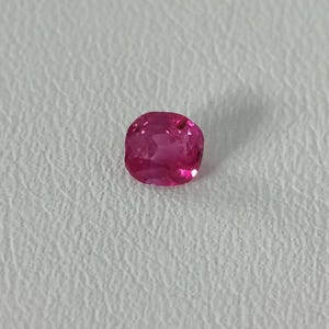 May include: A cushion-cut gemstone with a vibrant, saturated pink color. The stone features a faceted surface that reflects light, creating internal highlights and shadows across its translucent body, showcasing a clear, polished appearance.