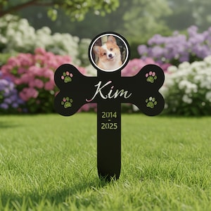 May include: A black pet memorial stake in the shape of a bone, with a photo of a dog in a circular frame. The name "Kim" is written in cursive, with the years 2014-2025 below. Green paw prints are on the bone.